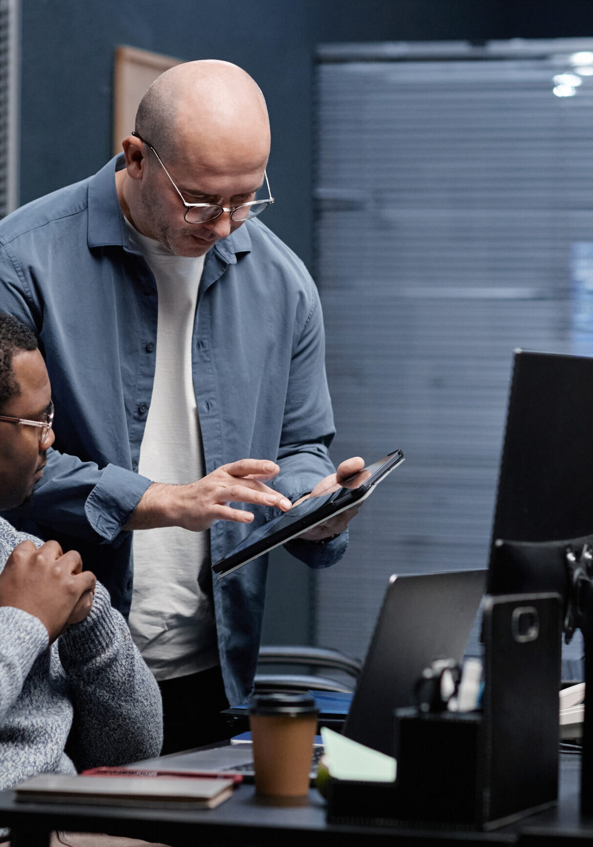 Middle aged Caucasian man showing digital tablet to young adult Black man in modern office, both collaborating on project while sitting and standing near computer monitors